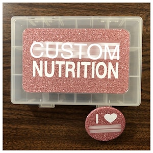 Puede incluir: Un recipiente de plástico transparente con 12 compartimentos, con una tapa de purpurina rosa con el texto "CUSTOM NUTRITION". También se incluye un botón redondo de purpurina rosa con un corazón blanco y el texto "I".