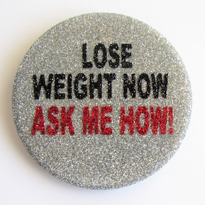 Puede incluir: Un botón de purpurina plateado con el texto "Lose Weight Now Ask Me How!" en letras negras y rojas.