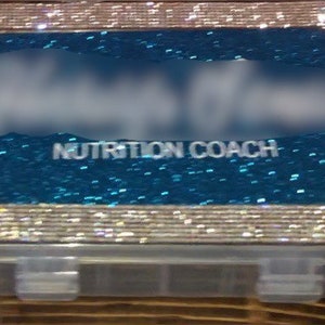 Puede incluir: Una caja de pastillas de plástico plateada y azul con un acabado brillante. La tapa tiene el texto "NUTRITION COACH" impreso.