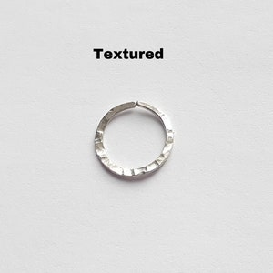 May include: A silver-coloured circular ring with a hammered, textured surface. The metal band features a small gap at the top, creating an open hoop design. The word Textured is printed in black text above the silver ring on a plain background.