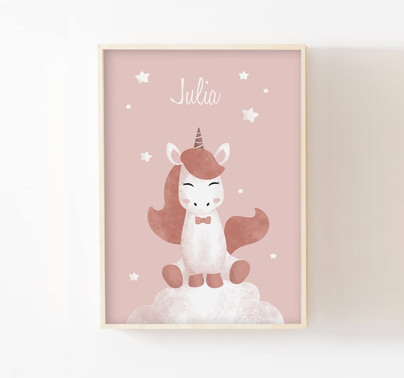 May include: A pink framed print featuring a white unicorn sitting on a cloud. The unicorn has a brown mane and tail, a gold horn, and a pink bow tie. The print also includes white stars and the name "Julia" in white text.