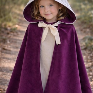 May include: A child's purple velvet cloak with a silver sequined hood and cream-coloured bow. The cloak is a deep shade of purple and has a soft, luxurious appearance. The hood is lined with a matching purple fabric.
