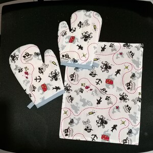 Könnte beinhalten: Ein weißes Küchenset aus Baumwolle mit Piratenmotiv. Das Set enthält zwei Ofenhandschuhe und ein passendes Geschirrtuch. Das Design zeigt Piratenschiffe, Schatztruhen, Anker und Tintenfisch-Illustrationen in Schwarz, Rot und Grau.