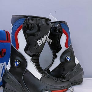 Puede incluir: Par de botas de moto negras y blancas con detalles en rojo y azul. Las botas presentan el logotipo de BMW y están diseñadas para la protección y el estilo. Las botas tienen un acabado brillante y protector.