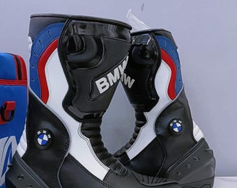 Puede incluir: Par de botas de moto negras y blancas con detalles en rojo y azul. Las botas presentan el logotipo de BMW y están diseñadas para la protección y el estilo. Las botas tienen un acabado brillante y protector.