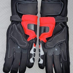 May include: A pair of black and red motorcycle gloves featuring reinforced knuckle protection, adjustable wrist straps with hook-and-loop closures, white accent panels on the fingers, and small circular logos on the back of the hands.