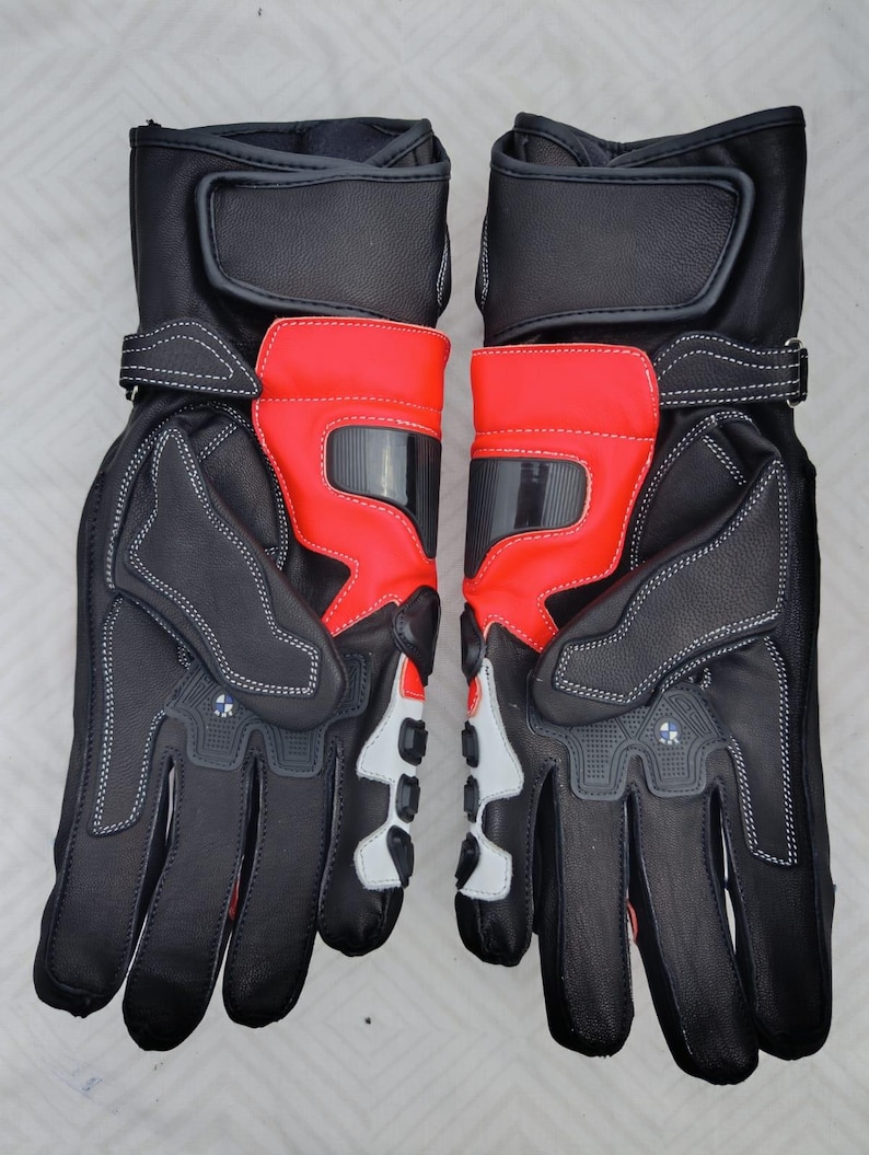 May include: A pair of black and red motorcycle gloves featuring reinforced knuckle protection, adjustable wrist straps with hook-and-loop closures, white accent panels on the fingers, and small circular logos on the back of the hands.