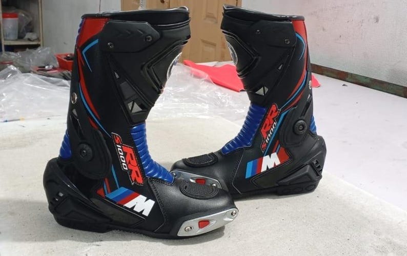 May include: A pair of black motorcycle boots featuring red, blue, and white accents. The boots display the text RR S1000 and the letter M on the side. They include protective panels, a blue accordion flex panel at the ankle, and a silver toe slider.