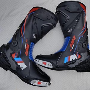 May include: A pair of black motorcycle racing boots featuring blue and red accents, with the text S 1000 RR and the M logo printed on the side. The boots include silver-toned toe sliders with red inserts and protective molded panels on the shins.