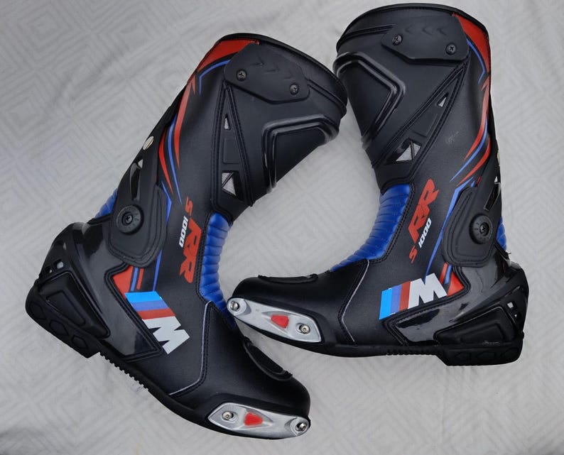 May include: A pair of black motorcycle racing boots featuring blue and red accents, with the text S 1000 RR and the M logo printed on the side. The boots include silver-toned toe sliders with red inserts and protective molded panels on the shins.