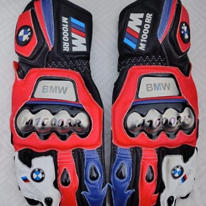 May include: A pair of black, red, white, and blue motorcycle gloves featuring the BMW M1000RR logo. The gloves include metallic knuckle protectors, branded patches on the cuffs, and circular BMW emblems on the wrist and finger areas.