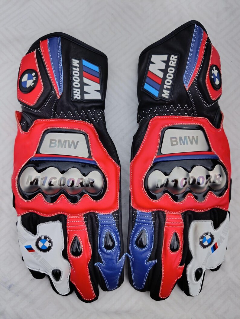 May include: A pair of black, red, white, and blue motorcycle gloves featuring the BMW M1000RR logo. The gloves include metallic knuckle protectors, branded patches on the cuffs, and circular BMW emblems on the wrist and finger areas.
