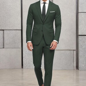 May include: A dark green suit with a white shirt and a dark tie. The suit jacket has a single button closure and a pocket on the left side. The man is wearing black shoes.