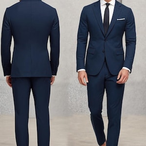 May include: A dark blue suit with a single-breasted jacket and matching trousers. The jacket has a pocket on the left side and a button fastening. The suit is worn by a person with brown hair and a white shirt with a dark blue tie.