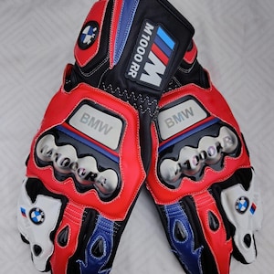 May include: A pair of black, red, and blue motorcycle gloves featuring the BMW logo and M1000 RR branding. The gloves include silver-toned knuckle protectors with embossed text, white accents on the fingers, and stitched detailing throughout.