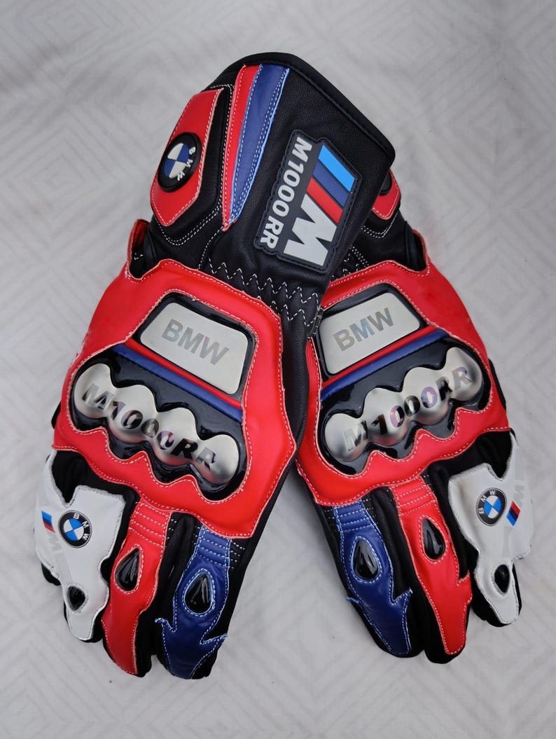 May include: A pair of black, red, and blue motorcycle gloves featuring the BMW logo and M1000 RR branding. The gloves include silver-toned knuckle protectors with embossed text, white accents on the fingers, and stitched detailing throughout.