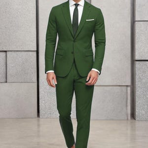 May include: A dark green suit with a white shirt and a dark green tie. The suit jacket has a single button closure and a pocket on the left side. The man is wearing black shoes.