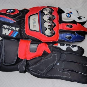 May include: A pair of black, red, white, and blue motorcycle gloves featuring the BMW M1000 RR logo. The gloves include protective knuckle guards, reinforced palm padding, adjustable wrist straps, and branded detailing on the cuffs and fingers.