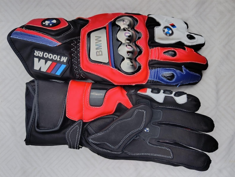 May include: A pair of black, red, white, and blue motorcycle gloves featuring the BMW M1000 RR logo. The gloves include protective knuckle guards, reinforced palm padding, adjustable wrist straps, and branded detailing on the cuffs and fingers.