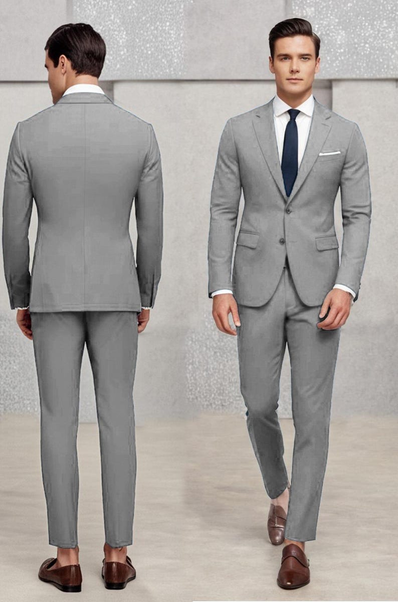 May include: A grey suit with a single-breasted jacket and matching trousers. The jacket has a pocket on the left side and a button fastening. The suit is worn by a person with brown hair and a white shirt with a dark blue tie.