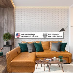 Op de afbeelding: Een beige lederen hoekbank met een chaise longue, voorzien van donkergroene en grijze sierkussens. Voor de bank staan twee houten salontafels. Een witte bakstenen muur vormt de achtergrond voor deze meubelopstelling in de ruimte.