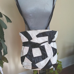 May include: A black and white patchwork leather shoulder bag. The bag is made from irregularly shaped black and white leather pieces. It features a drawstring closure and black shoulder straps. The bag is displayed on a mannequin.