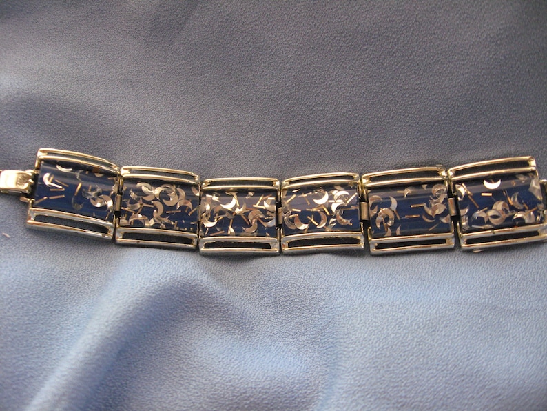 May include: A gold and blue bracelet with a repeating pattern of silver glitter. The bracelet is made of metal and has a hinged clasp.