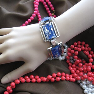 May include: A silver and blue bracelet with a floral design. The bracelet is on a mannequin's wrist and surrounded by pink and white beads.
