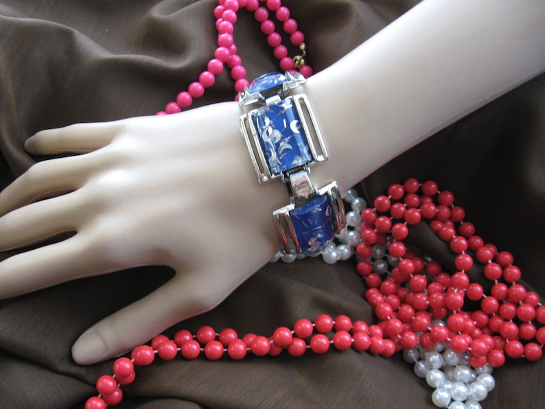 May include: A silver and blue bracelet with a floral design. The bracelet is on a mannequin's wrist and surrounded by pink and white beads.