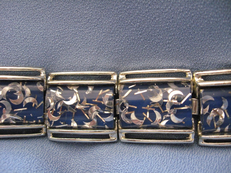 May include: A silver bracelet with a blue background and silver glitter. The glitter is shaped like crescent moons.