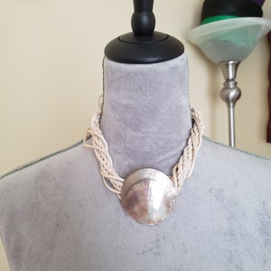 以下が含まれることがあります： 白いロープのネックレスに、大きな虹色の貝殻のペンダントが付いている。