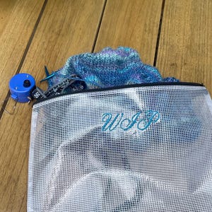 May include: A clear, mesh-textured zipper pouch with the letters WIP embroidered in blue thread. Inside the pouch are blue and purple variegated yarn, knitting needles, and a blue retractable measuring tape showing inches.
