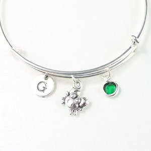 May include: Silver bangle bracelet with a charm of a crab, a green gemstone charm, and a charm with the letter G.
