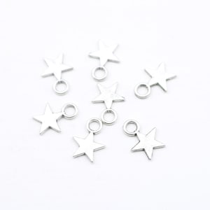 May include: A group of eight silver star-shaped charms with a jump ring attached to each star. The charms are scattered on a white surface.
