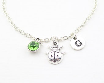 Puede incluir: Pulsera de cadena plateada con un dije de cristal verde, un dije de mariquita y un dije con la letra G.