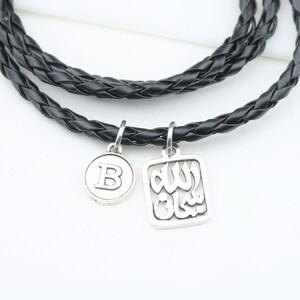 Op de afbeelding: Zwarte gevlochten leren ketting met twee zilveren hangers. Eén hanger is een ronde schijf met de letter "B", de andere is een vierkante hanger met Arabisch schrift. De ketting wordt gepresenteerd op een wit oppervlak.