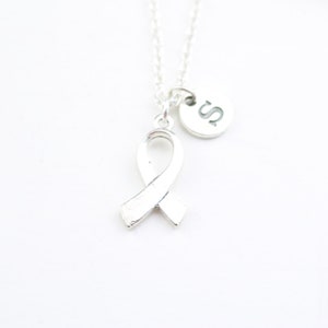 Op de afbeelding: Zilverkleurige ketting met een lintvormige hanger en een kleine ronde bedel met de letter "S". Het lint is een symbool van bewustwording. De ketting hangt tegen een witte achtergrond.