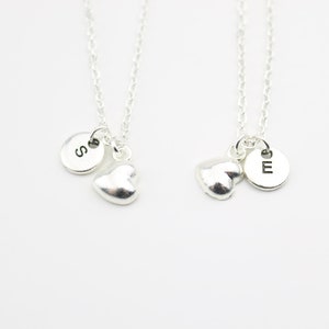 May include: Two silver necklaces with heart charms and initial discs. The necklace on the left has a disc with the letter 'S' and the necklace on the right has a disc with the letter 'E'.