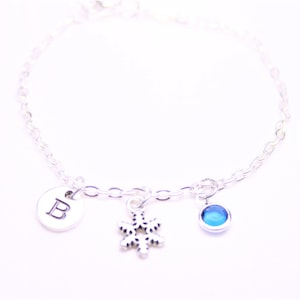 May include: A silver chain bracelet with a snowflake charm, a blue gemstone charm, and a silver charm with the letter "B" engraved on it.