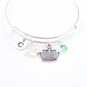 May include: Silver charm bracelet with a crown charm, a letter C charm, a number 21 charm, and a green gemstone charm.