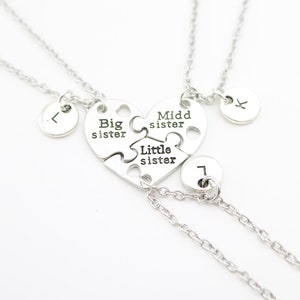 May include: Three silver necklaces with heart-shaped puzzle pieces that say "Big sister", "Middle sister", and "Little sister". Each necklace has a small round charm with a letter on it.