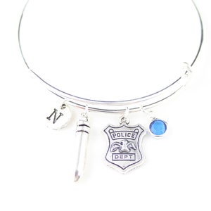 Puede incluir: Pulsera de plata con un dije de placa de policía, un dije de letra N, un dije de plata que parece un micrófono y un dije de cristal azul.