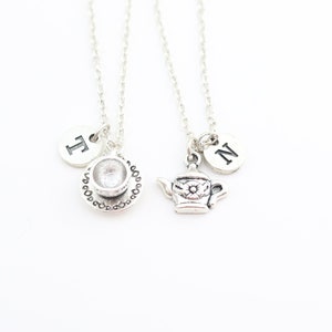 Op de afbeelding: Twee zilveren kettingen met bedels. Eén ketting heeft een theekopje-bedel en een bedel met de letter "T". De andere ketting heeft een theepot-bedel en een bedel met de letter "N".