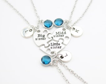 May include: Three silver necklaces with puzzle piece heart pendants that say "Big sister", "Middle sister", and "Little sister". Each necklace has a small silver disc with a letter on it and a blue gemstone.