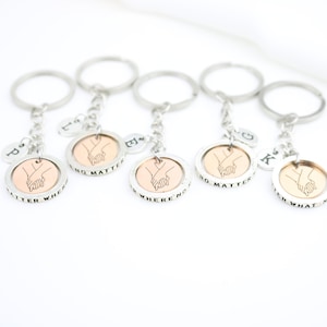 May include: Five silver keychains with a copper-colored disc featuring a line drawing of two hands holding each other. Each disc has a different inscription: "No matter where", "No matter when", "No matter what", "No matter how", and "No matter who". Each keychain also has a small silver charm with a letter on it.