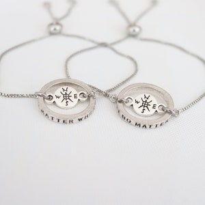 May include: Two silver friendship bracelets with adjustable cords. Each bracelet features a compass charm with the words "No matter" and "Matter what" inscribed around the circle.