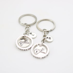 May include: Two silver keychains with a circle charm featuring a pinky promise hand gesture and the text "Where there is no" and "That no matter". Each keychain has a small silver charm with the letters "L" and "M" on them.