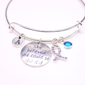 May include: A silver charm bracelet with a charm that says "She believed she could so she did". The bracelet also has a charm with the letter A and a blue crystal charm.