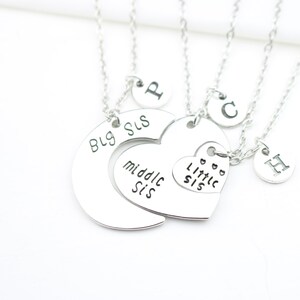 May include: Three silver necklaces with heart and circle pendants. The pendants have the words "Big Sis", "Middle Sis", and "Little Sis" engraved on them. Each necklace also has a small circle pendant with a letter engraved on it.