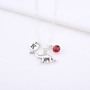 Pode incluir: Colar de prata com um pingente de dinossauro, um pingente de cristal vermelho e um pingente de prata com a letra K.